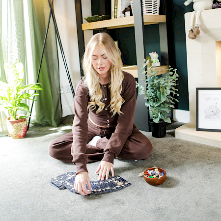 A woman sitting in a cross-legged meditative pose on a yoga mat in a sunlit forest.