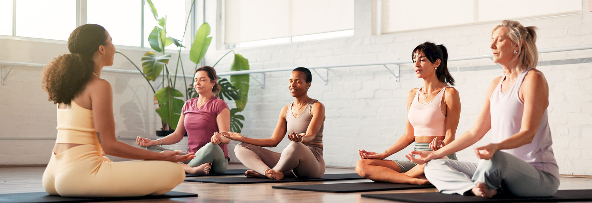 A group of women sitting in a circle on yoga mats, practicing mindfulness meditation in a bright studio.