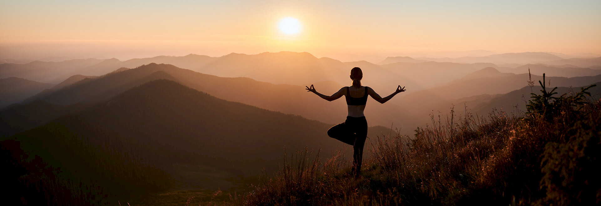 Silhouette of a woman practicing a tree yoga pose on a mountain ridge at sunset.