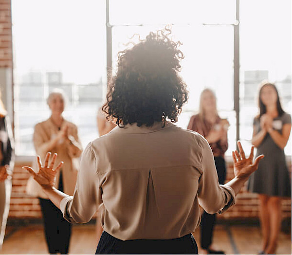 Rear view of a woman with curly hair gesturing with her hands while speaking to a group in a sunlit loft.