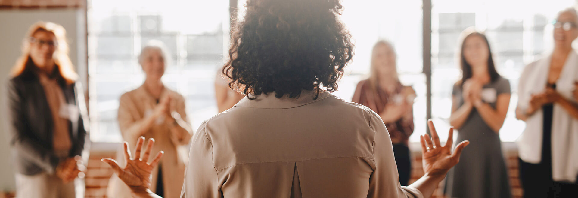 A speaker with curly hair stands before an audience in a bright, brick-walled studio as they applaud.