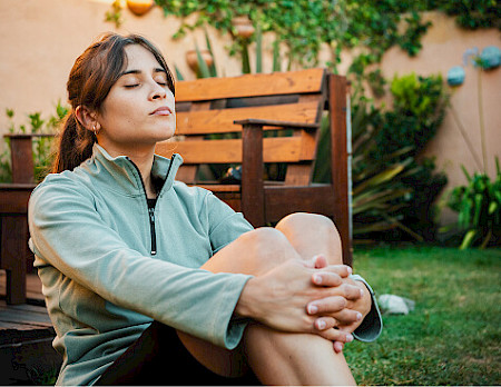 Young woman meditating peacefully while sitting on lawn near wooden bench in outdoor patio