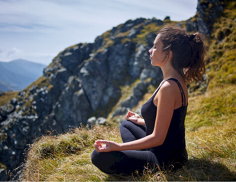 Woman meditating in lotus position on a grassy mountainside with rocky peaks in the background.
