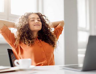 Relaxed woman in orange blouse taking a satisfying break at her workplace with laptop and coffee cup nearby.