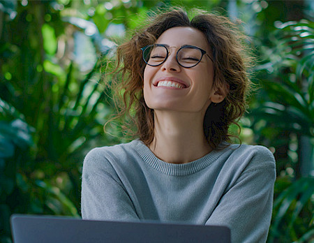 Woman with curly hair and glasses smiling joyfully while working on laptop surrounded by green plants.